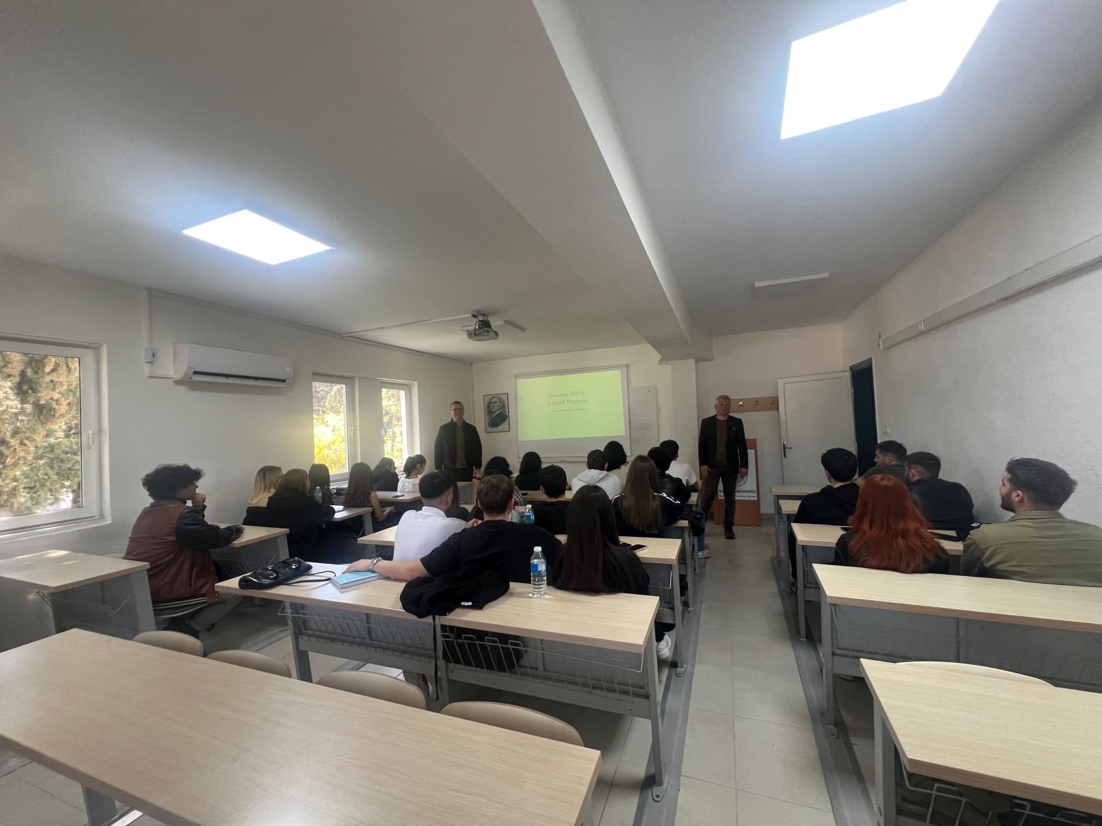 Sınıf ortamında gerçekleştirilen bilgilendirme toplantısında öğrenciler sıralarda otururken, iki konuşmacı projeksiyon perdesinin önünde ayakta sunum yapıyor. Aydınlık derslikte projeksiyon cihazı, klima ve pencereler görülüyor.